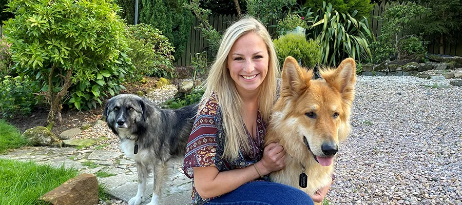Woman smiling at camera crouched down with two dogs in a garden.